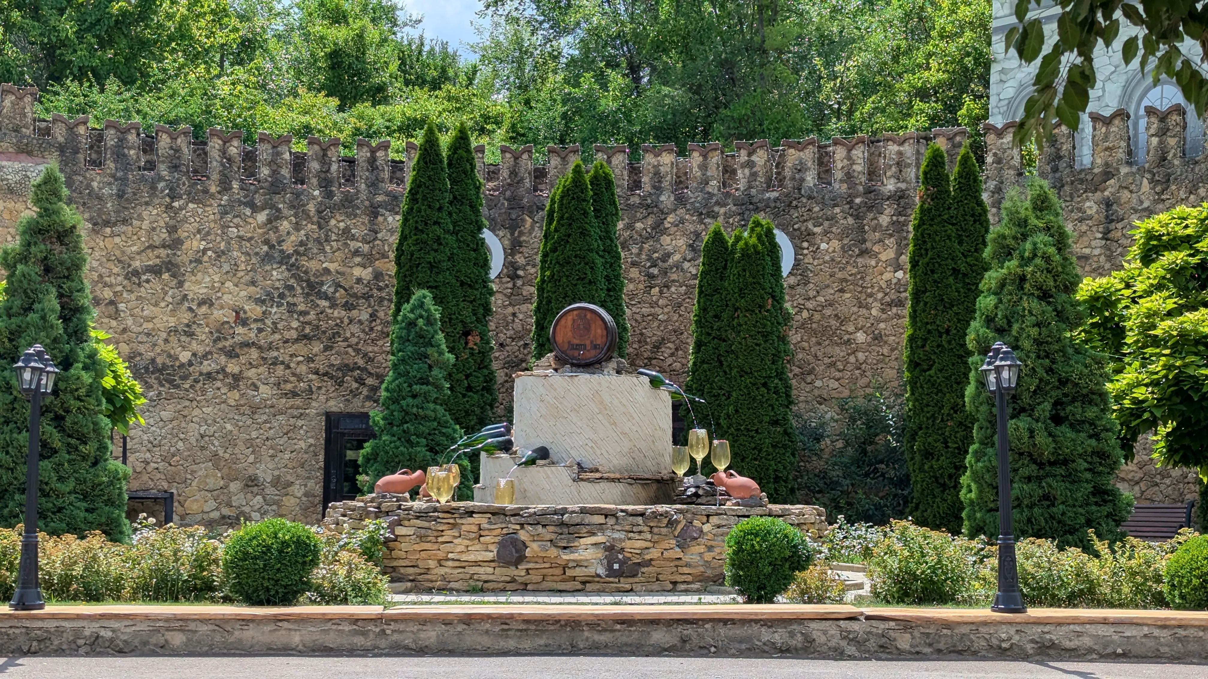 Fontanna z wielkimi kieliszkami udająca białe wino, w tle kamienny mur i
drzewa