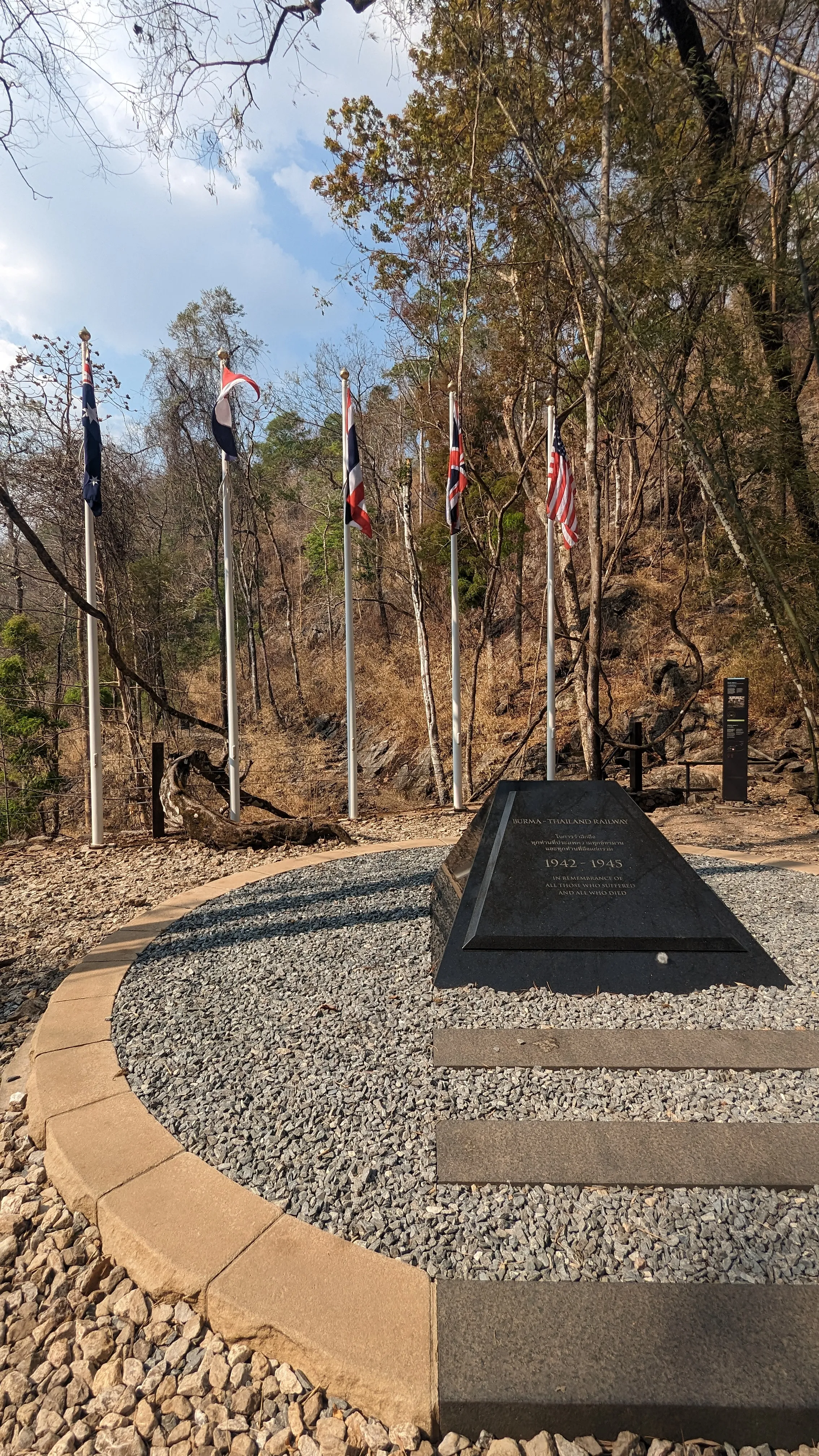 Granitowy stożek z datami i pamiątkową inskrypcją po tajsku i angielsku, otoczony półkolem
masztów z flagami, z przodu ścieżka z kamiennymi płytami przypominającymi podkłady
kolejowe