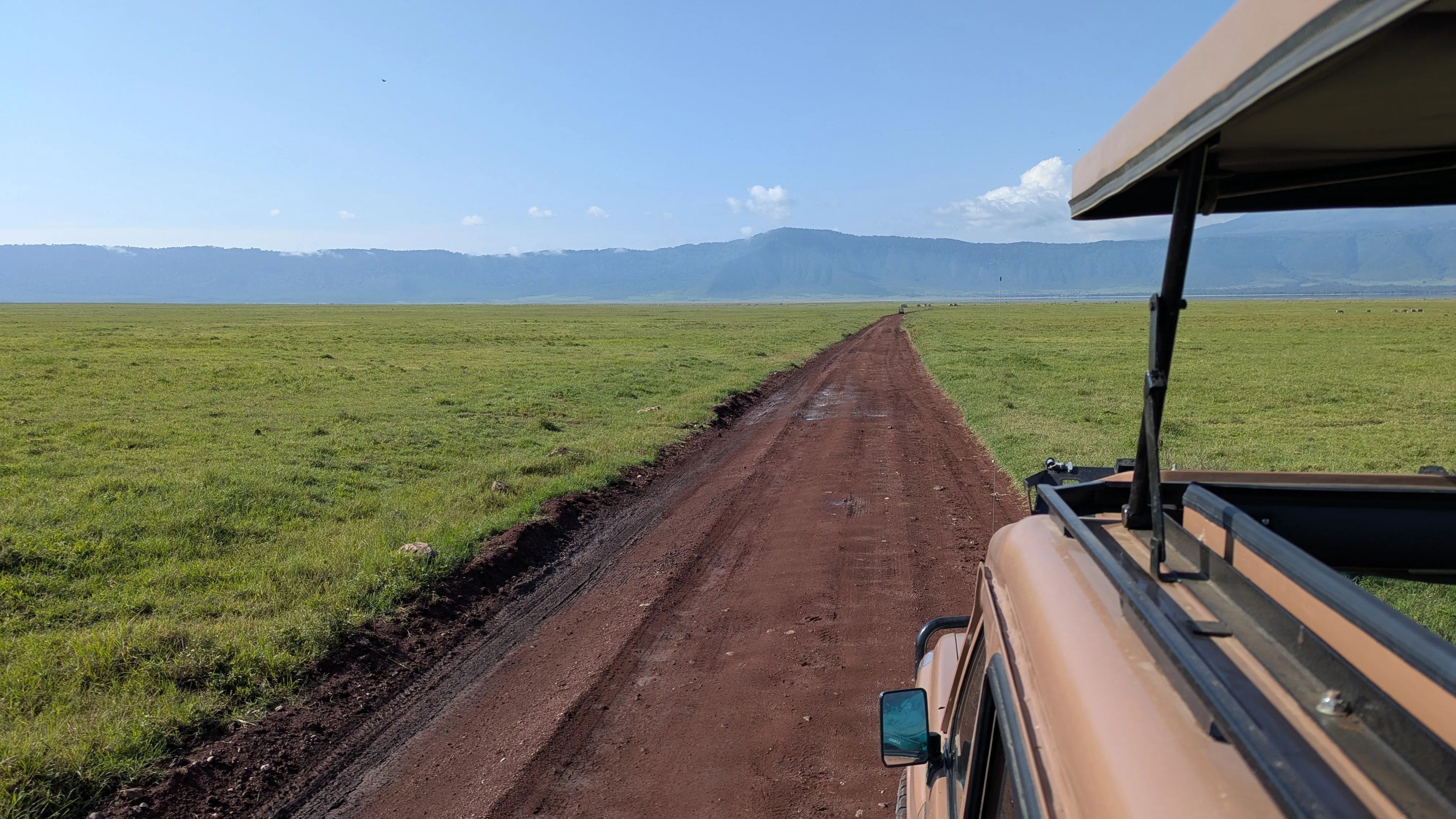 Zieloną trawę przecina rudoczerwona droga gruntowa, w prawym brzegu widać samochód z
podniesionym dachem, z którego jest robione zdjęcie. W tle
wzgórza
